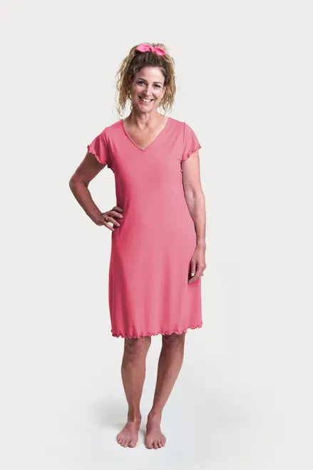 A woman with curly hair in a ponytail, barefoot against a white background, wears the Goodnighties Flutter Sleeve Knee Length Gown in pink with a matching scrunchie.