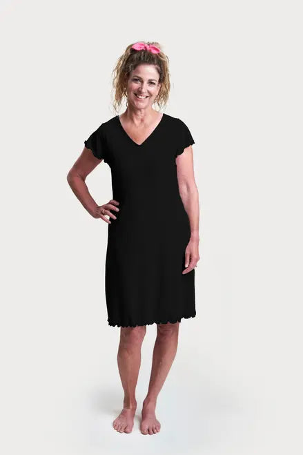 A smiling woman with curly hair in a pink scrunchie stands barefoot against a white background, wearing Goodnighties’ Flutter Sleeve Knee Length Gown—a stylish, black V-neck nightgown featuring 37.5 thermoregulating technology.