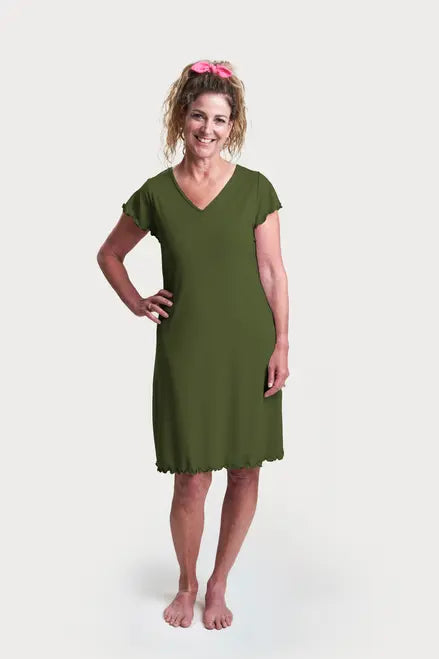 A smiling woman with curly hair in a pink scrunchie wears the Goodnighties Flutter Sleeve Knee Length Gown, an olive green, thermoregulating nightgown, standing barefoot against a plain white background.