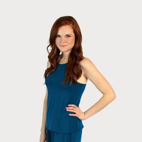 A young woman with long red hair stands smiling with one hand on her hip, modeling the sleeveless blue Lace Back Halter Top by Goodnighties against a plain light gray background.