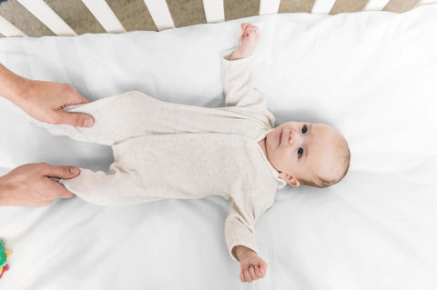 A baby lies calmly in a crib while an adult gently holds its legs, resting on the Soft Cotton Baby Bedding Crib Sheet by American Blossom Linens in a soothing white tone.