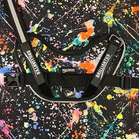 A BrilliantK9 Ergonomic Dog Harness for XLarge Dogs (80 lbs and up), in black with reflective strips and an adjustable chest strap, is shown on a multicolored splatter-paint background with bold orange, green, yellow, pink, and blue accents.