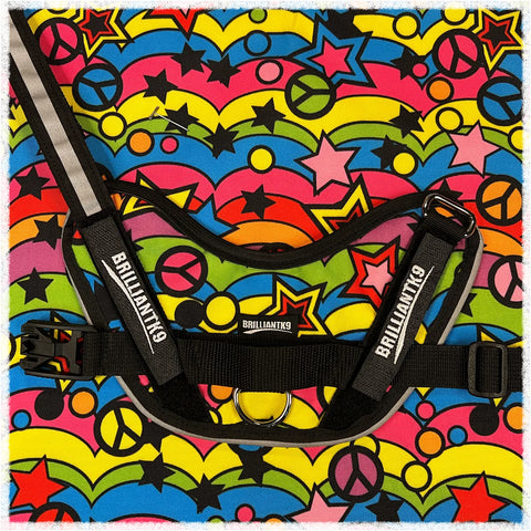 A BrilliantK9 Ergonomic Dog Harness for large dogs (50–80 lbs) rests on a bright, patterned background with stars, rainbows, peace signs, and hearts in yellow, pink, blue, green, orange, and black.