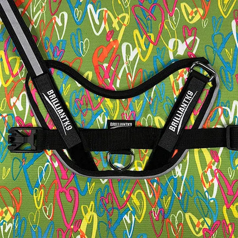 The BrilliantK9 Ergonomic Dog Harness for large dogs (50–80 lbs) stands out on a green background with colorful, overlapping heart doodles in blue, red, yellow, and pink.