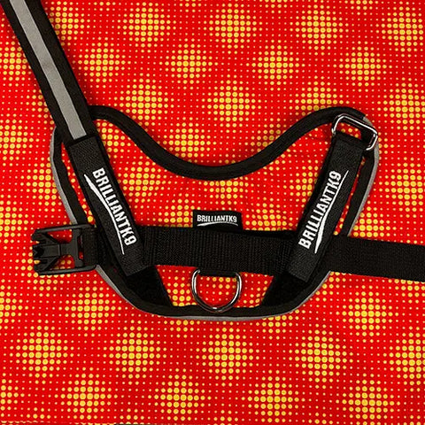 A BrilliantK9 Ergonomic Dog Harness for large dogs (50-80 lbs) lies on a red and yellow dotted background. Black, it features an adjustable chest strap, reflective strip, buckle, and a center metal ring.