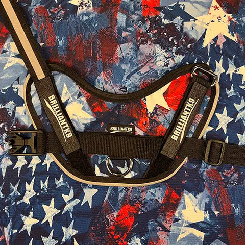A BrilliantK9 Ergonomic Dog Harness for large dogs (50–80 lbs), in black with reflective stripes and visible branding, rests on a red, white, and blue abstract star-patterned fabric. It features an adjustable chest strap for comfort and durability.
