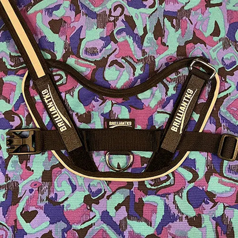 A BrilliantK9 Ergonomic Dog Harness for little dogs (10lbs & under), with BRILLIANTK9 on the straps, rests on a vibrant blue, purple, and pink abstract-patterned fabric.