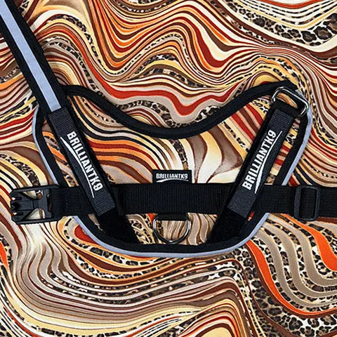 A BrilliantK9 Ergonomic Dog Harness for little dogs 10lbs & under, in black with reflective strips and the BrilliantK9 logo, rests on wavy-patterned orange, brown, and cream fabric.