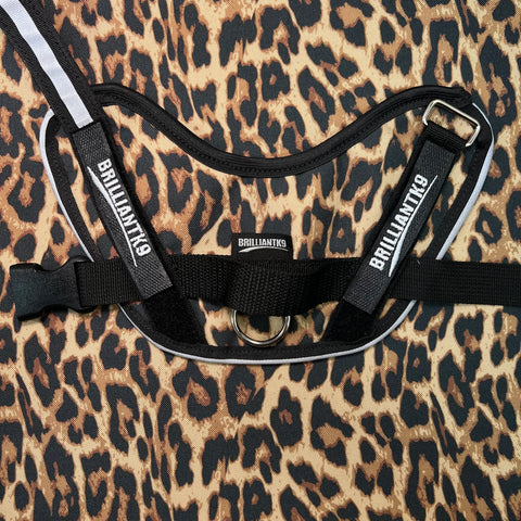 A black, reflective BrilliantK9 Medium Dog Harness – Ergonomic No-Pull Comfort (20-45 lbs) by BrilliantK9 is shown on a leopard print fabric. The harness features branded labels and a sturdy central metal ring. Made in USA.