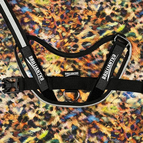 A black BrilliantK9 Ergonomic Dog Harness for medium dogs (20–45 lbs) is displayed with reflective stripes on a colorful animal-print fabric background.