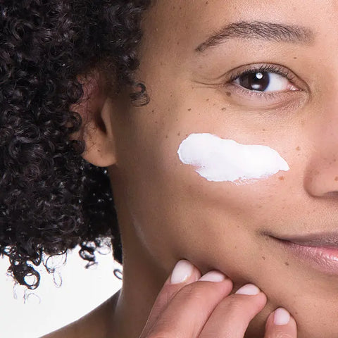 Close-up of a person with curly hair applying Open Formula SPF 30 Sheer Moisturizer to their cheek, smiling slightly, with a focus on their eye and fingers touching their face to help prevent premature aging.