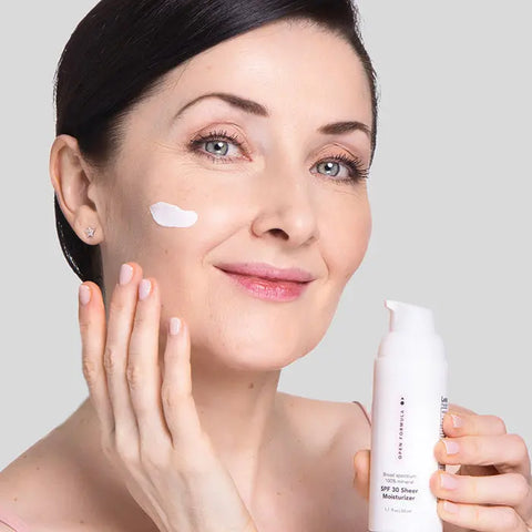 A fair-skinned woman smiles as she applies Open Formula SPF 30 Sheer Moisturizer to her cheek, holding the white bottle against a light gray background.