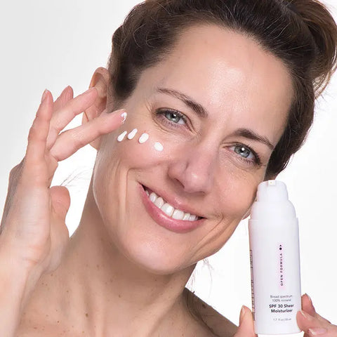 A woman with clear skin and brown hair applies dots of Open Formula SPF 30 Sheer Moisturizer to her cheek, holding the white bottle in one hand, ready to protect against sun damage and premature aging.