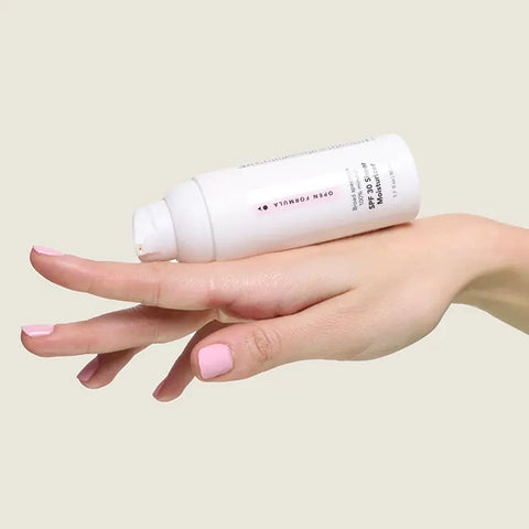 A hand with light pink nail polish holds the Open Formula SPF 30 Sheer Moisturizer bottle horizontally against a light beige background.