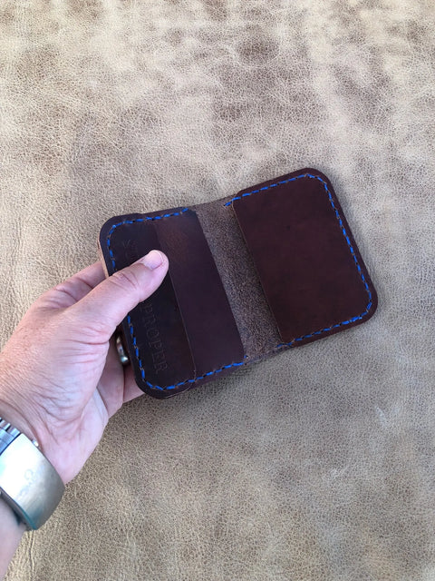 A hand holds an open Sully + Proper Supply Buffalo Nickel Card Wallet in Dark Oil Brown with blue stitching, showing two empty card slots. The American-made minimalist wallet rests on a textured light brown background.