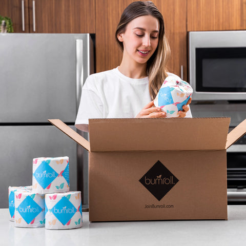 A woman smiles in a modern kitchen as she unpacks a Bumroll box filled with Bumroll Premium Toilet Paper, which comes in colorful wraps and helps plant trees in the USA.