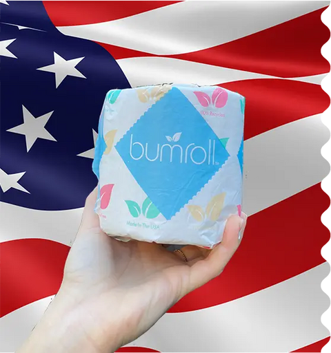 A hand holds a roll of Bumroll Premium Toilet Paper, which plants trees in the USA, in front of an American flag. The packaging features colored leaves and a Made in the USA label, showing Bumroll’s eco-friendly commitment.