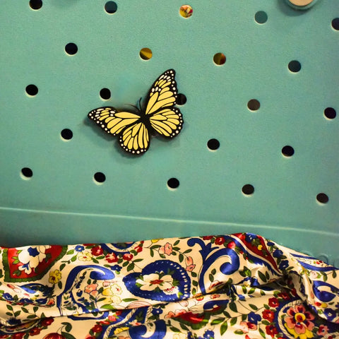 The FRESHe Baglets Bug Charm by Chatelet Manufacturing features a yellow and black butterfly charm on a blue tote, ideal as a decorative accessory for Bogg Bags, Simply Southern, and other totes with colorful floral patterns.