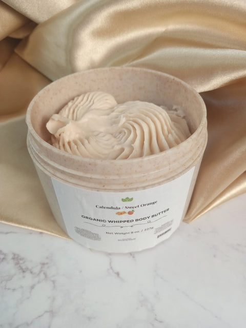 An open jar of Forev'Herbs Calendula Sweet Orange Organic Whipped Body Butter, with a creamy swirl texture, rests on marble beside gold satin. Its label highlights nourishment for dry, sensitive skin with a fresh calendula orange scent.