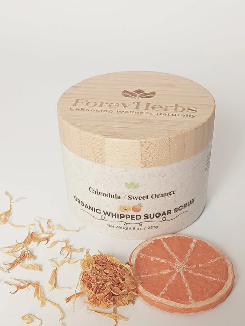 A jar of Forev'Herbs Calendula Sweet Orange Organic Whipped Sugar Scrub with a bamboo lid is displayed beside dried calendula petals and an orange slice, showcasing the sweet orange essence of this exfoliating scrub on a white background.