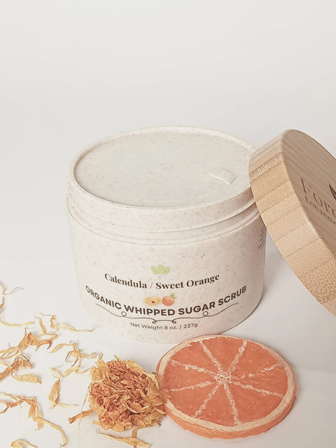 A jar of Forev'Herbs Calendula Sweet Orange Organic Whipped Sugar Scrub sits open with its bamboo lid nearby, surrounded by dried petals and a dried orange slice on a white surface, highlighting its exfoliating and sweet orange essence.