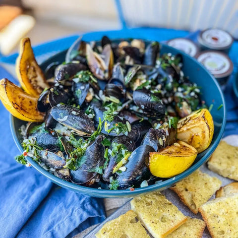 A blue bowl of cooked mussels garnished with herbs and grilled lemon sits on a blue cloth next to bread made with Gustus Vitae’s California Seasonings Gift Set – Tastes of California artisanal spice blends. Sauce containers are blurred in the background.