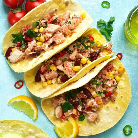 Three soft tacos filled with shredded chicken, corn, beans, tomatoes, herbs, and chili are served on a light blue surface with lemon slices and cherry tomatoes, enhanced by Gustus Vitae’s California Seasonings Gift Set artisanal spice blends.