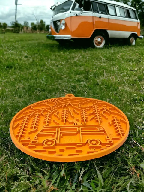 The SodaPup Camp Design EMat Enrichment Lick Mat With Suction Cups is shown on grass with an orange ornament featuring a camper van and forest, while a real vintage van appears blurred in the background.