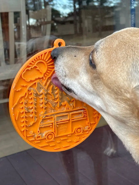 A dog licks the SodaPup Camp Design EMat Enrichment Lick Mat with suction cups, featuring trees, mountains, a van, and camp embossed on its orange round surface. The mat is stuck to a glass door.