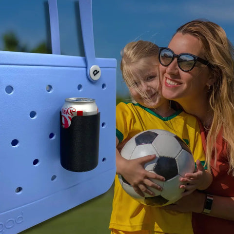 A smiling woman in sunglasses hugs a young girl with a soccer ball. In the foreground, a Chatelet Manufacturing FRESHe BAGLETS Can Drink & Water Bottle Holder Charm Accessory is attached to a tote bag next to a Diet Coke can. Sunny outdoor setting in the background.