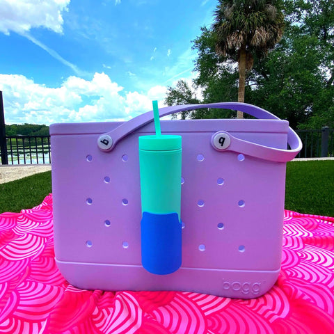 A lavender Rubber Tote Bag holds a teal cup in a Chatelet Manufacturing FRESHe BAGLETS Can Drink & Water Bottle Holder Charm, set on a pink blanket outdoors with trees, a fence, and blue skies creating a relaxing scene.