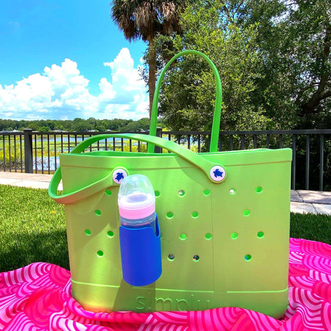 A Chatelet Manufacturing FRESHe BAGLETS Can Drink & Water Bottle Holder Charm Accessory hangs from a green rubber tote bag with grommet holes and googly eyes, resting on a pink blanket outdoors by trees, a lake, and blue sky.