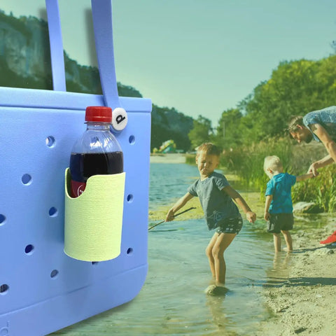 A close-up of a FRESHe BAGLETS Can Drink & Water Bottle Holder Charm Accessory by Chatelet Manufacturing, attached to a light blue tote bag, with a man and two children playing by the riverside on a sunny day.
