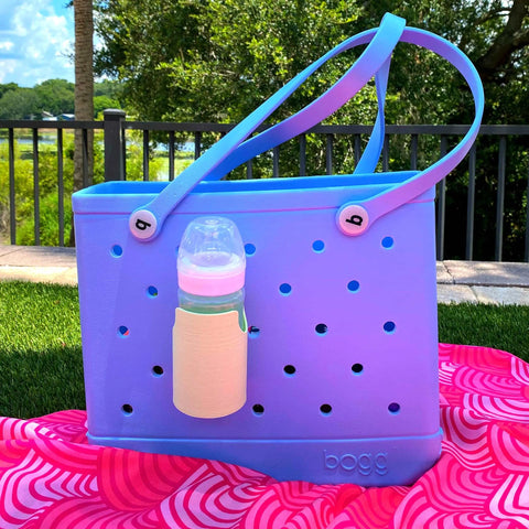 Outdoors, a blue and purple rubber tote sits on a pink swirled-pattern towel. A baby bottle in the Chatelet Manufacturing FRESHe BAGLETS bottle holder charm accessory is attached to the bag, keeping drinks handy amid grass, trees, and a fence.