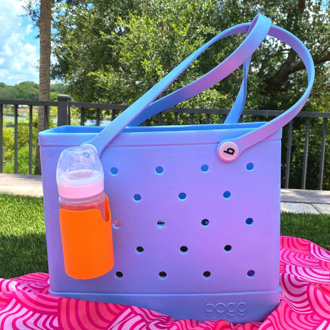 Outdoors, a blue tote bag with round holes sits on a pink towel. Attached to its handle is an orange FRESHe BAGLETS Can Drink & Water Bottle Holder Charm Accessory by Chatelet Manufacturing, compatible with Bogg Bags. Trees and a black fence are in the background.