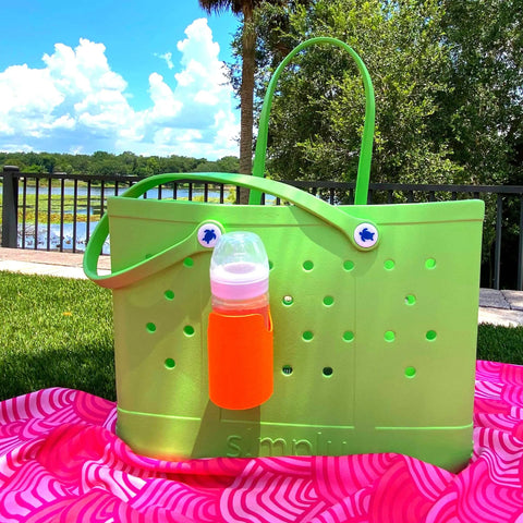Outdoors on a pink and white blanket, a large light green Rubber Tote Bag with blue and white eye decorations features a FRESHe BAGLETS Can Drink & Water Bottle Holder Charm Accessory by Chatelet Manufacturing hanging from the front. Trees and water are in the background.