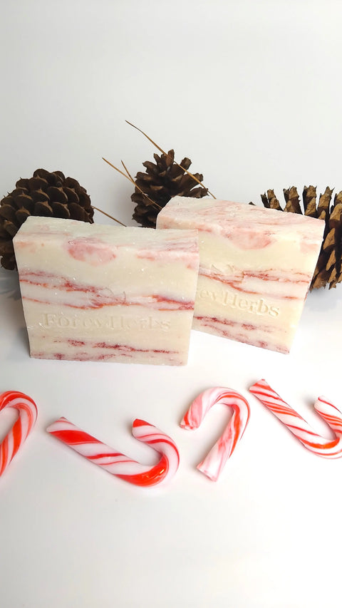 Two bars of Forev'Herbs Candy Cane Handmade Soap, marbled pink and white with embossed branding, are displayed before pinecones. Four mini red and white candy canes complete this festive holiday skincare scene.
