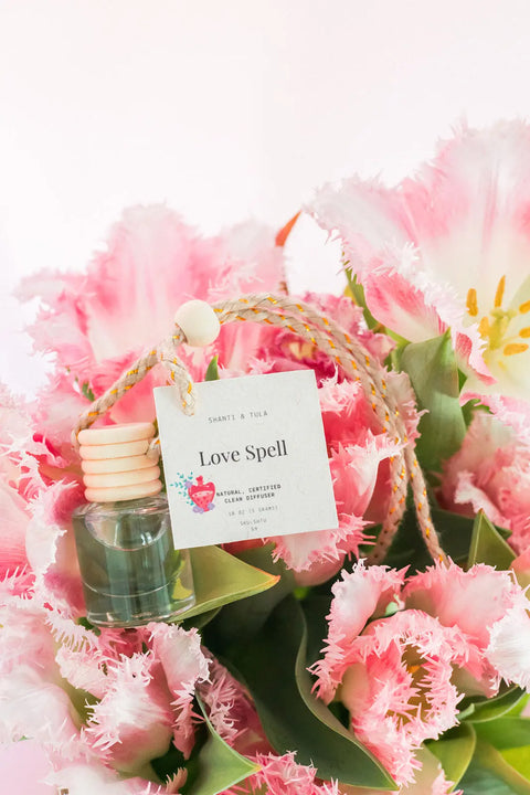 The Shanti + Tula Car Diffuser & Refill Set is a premium hanging air freshener made in the USA, featuring pink fringed tulips, a “Love Spell” labeled bottle with heart graphic, wooden bead detail, and long-lasting scents—perfect for your car.