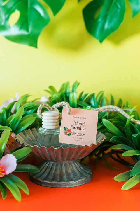 A Shanti + Tula Car Diffuser & Refill Set, a long-lasting hanging air freshener made in the USA, rests on a metal stand among faux greenery and pink flowers, with its bottle displayed against a vibrant yellow background.