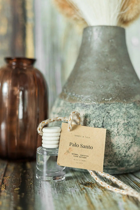 A Shanti + Tula Car Diffuser & Refill Set—Long-Lasting Hanging Air Freshener—sits on wood beside brown and gray vases with dried flowers. The glass bottle has a wooden cap, rope, and Palo Santo label. Made in USA.