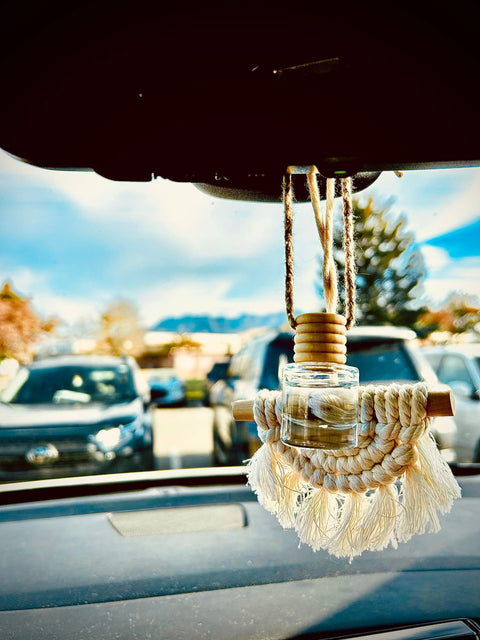 A Shanti + Tula Car Diffuser & Refill Set, a long-lasting hanging air freshener made in the USA, with twine and macramé fringe, hangs from a rearview mirror as parked cars and a cloudy sky can be seen through the windshield.