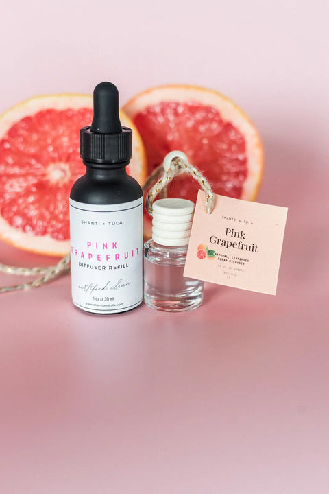 A dropper bottle and vial labeled Pink Grapefruit, alongside a halved grapefruit on a pink surface, highlight the Shanti + Tula Car Diffuser & Refill Set—premium, long-lasting hanging air fresheners made in the USA.