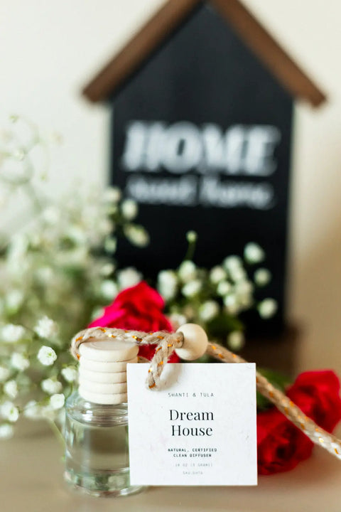 A compact Shanti + Tula Car Diffuser & Refill Set—long-lasting and made in the USA—features a small glass bottle with rope and wooden cap labeled Dream House, surrounded by red and white flowers before a blurred HOME sweet Home sign.