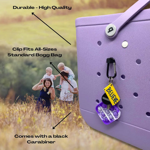 A purple rubber tote bag features the BAGLETS Carabiner & Plug Keys Holder Charm by Chatelet Manufacturing, a durable USA-made accessory for securing keys or valuables. Attached are a mini pop-it toy and yellow BELIEVE tag. A family plays in the field behind.