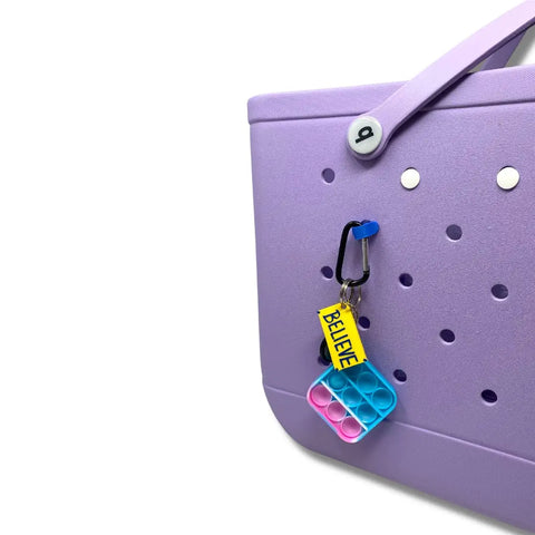 A light purple Bogg Bag with holes, a purple strap, and a Chatelet Manufacturing BAGLETS Carabiner & Plug Keys Holder Charm accessory—yellow “BELIEVE” tag and blue/pink pop-it toy attached—compatible with rubber tote bags.