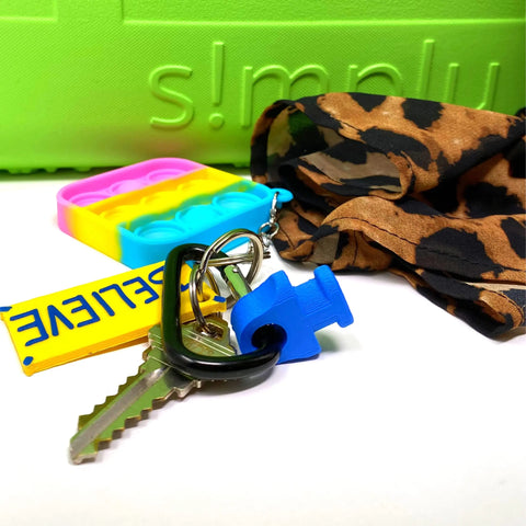 A set of keys with a yellow BELIEVE keychain, blue puzzle piece charm, pop-it toy, and leopard-print scarf rests on a bright green surface—featuring the BAGLETS Carabiner & Plug Keys Holder by Chatelet Manufacturing for rubber tote bags.