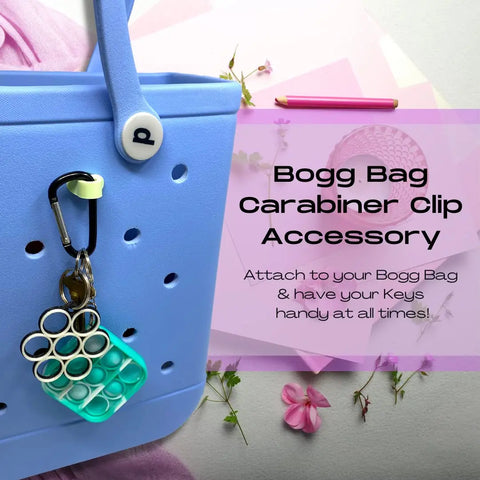 A blue rubber tote bag holds a small pop-it keychain with the Chatelet Manufacturing BAGLETS Carabiner & Plug Keys Holder Charm Accessory—keeping keys secure and handy. Pink papers, pencils, and a pink flower are nearby.