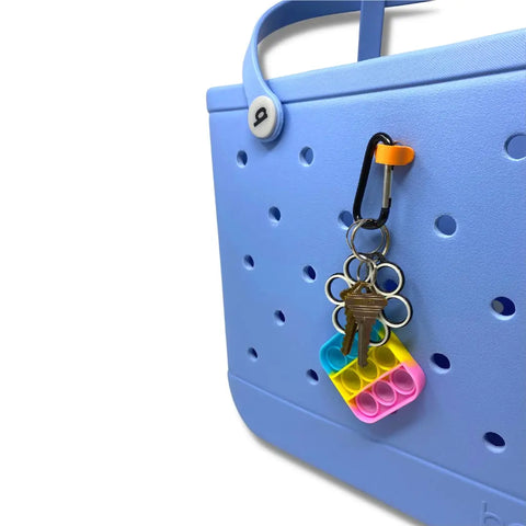 The BAGLETS Carabiner & Plug Keys Holder Charm Accessory by Chatelet Manufacturing secures keys or valuables to rubber tote bags and adds a colorful touch, shown here with a flower-shaped pop-it keychain on a light blue bag. Made in the USA.