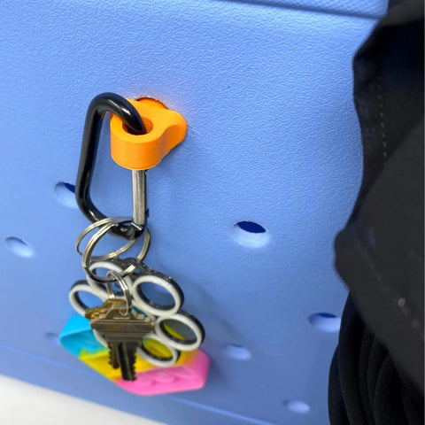 A set of keys with a colorful fidget keychain and the Chatelet Manufacturing BAGLETS Carabiner & Plug Keys Holder hangs on an orange hook on a blue pegboard. A black strap, likely from a rubber tote bag accessory, is visible to the right.