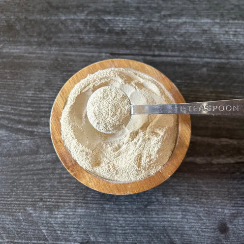 A metal teaspoon of light beige Oceans Balance Sea Moss Powder rests on a wooden bowl with more powder atop a dark surface—an essential ingredient known for supporting thyroid health.